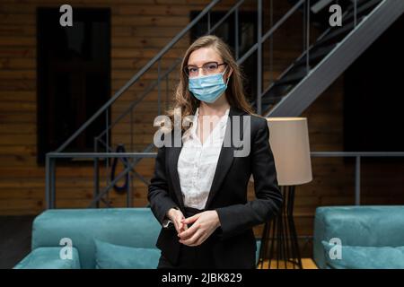 Eine junge Frau in einem schwarzen Business-Anzug sieht die Kamera mit einer medizinischen Schutzmaske im Gesicht an Stockfoto