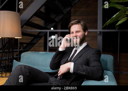 Ein männlicher Geschäftsmann sitzt in einem stilvollen Restaurant, entspannt in einem Geschäftsanzug und nutzt das Telefon, um zu kommunizieren Stockfoto