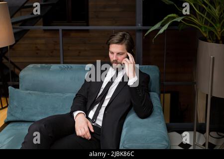 Ein männlicher Geschäftsmann sitzt in einem stilvollen Restaurant, entspannt in einem Geschäftsanzug und nutzt das Telefon, um zu kommunizieren Stockfoto