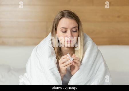 Junge Frau, die zu Hause auf dem Bett sitzt, bedeckt mit einer Decke, krank, den Kopf haltend, eine laufende Nase hat, niest, allergiert, fühlt sich schlecht an Stockfoto