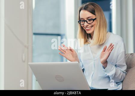 Schöne blonde Frau in legerer Kleidung sitzt auf einem Sofa zu Hause und nutzt einen Laptop für die Videokommunikation mit Freunden und Familie Stockfoto