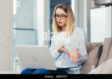 Schöne blonde Frau in legerer Kleidung sitzt auf einem Sofa zu Hause und nutzt einen Laptop für die Videokommunikation mit Freunden und Familie Stockfoto