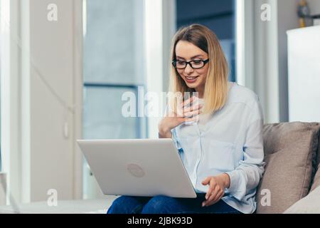 Schöne blonde Frau in legerer Kleidung sitzt auf einem Sofa zu Hause und nutzt einen Laptop für die Videokommunikation mit Freunden und Familie Stockfoto