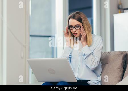 Schöne blonde Frau in legerer Kleidung sitzt auf einem Sofa zu Hause und nutzt einen Laptop für die Videokommunikation mit Freunden und Familie Stockfoto