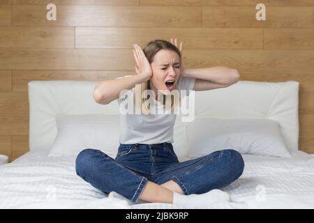 Wütende junge Frau, die zu Hause in einem Hotelzimmer auf dem Bett sitzt, bedeckt ihre Ohren mit den Händen vor dem lauten Lärm der Nachbarn, Reparaturen, Lärm von Th Stockfoto