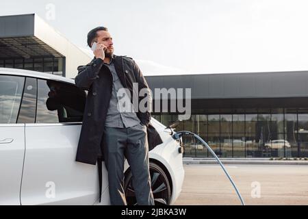 Ein eleganter Geschäftsmann, der bei Sonnenuntergang ein neues weißes Elektroauto auf dem Parkplatz des Handelszentrums lädt Stockfoto