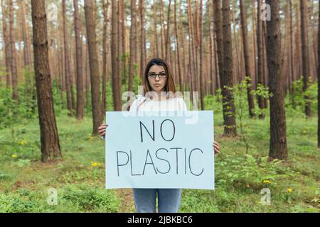 Junge Aktivistin, die ohne Plastikplakat im Wald steht, kämpft ehrenamtlich mit der Waldverschmutzung Stockfoto