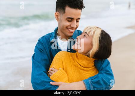 Multirassisches junges Paar, das am Strand lächelt und umarmt Stockfoto