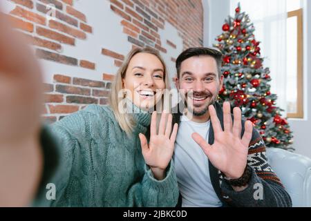 Frau und Mann zu Hause mit Blick auf Smartphone-Kamera begrüßen Freunde mit Urlaub, junge Familie während Neujahr und Weihnachten Stockfoto