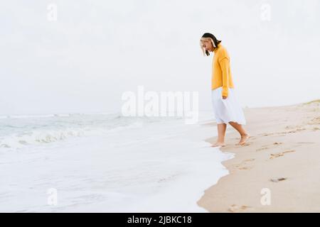 Junge asiatische Frau trägt Pullover lächelnd während des Spazierens am Strand Stockfoto