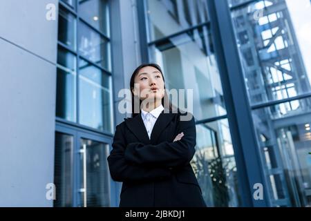 Erfolgreiche Geschäftsfrau nachdenklich und ernst mit gekreuzten Armen, außerhalb des Büros, asiatische Frau, die Kamera anschaut Stockfoto