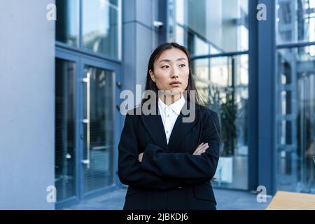 Erfolgreiche Geschäftsfrau nachdenklich und ernst mit gekreuzten Armen, außerhalb des Büros, asiatische Frau, die Kamera anschaut Stockfoto