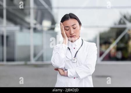 Junge asiatische Ärztin in weißem medizinischen Kleid in der Nähe der Klinik, müde depressiv, nach harten Arbeitstag depressiv überarbeitet Stockfoto