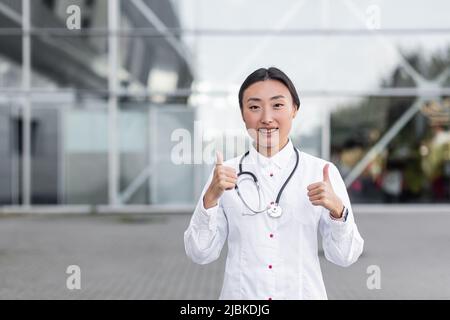 Ärztin lächelt und schaut in die Kamera, hält die Daumen hoch, positive asiatische Kinderärztin in der Nähe der Klinik Stockfoto