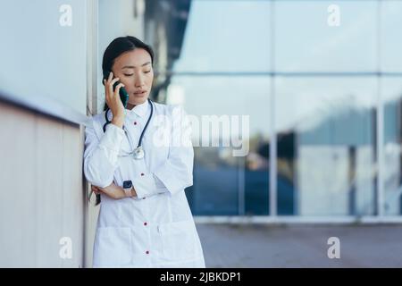 Traurige und frustrierte Ärztin, die in der Nähe der Klinik telefoniert, asiatisch depressiv und nach der Arbeit müde Stockfoto