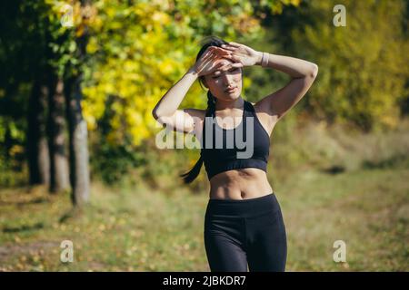 Eine Athletin läuft im Park, hat starke Kopfschmerzen, eine asiatische Frau hält ihre Hände hinter ihrem Kopf Stockfoto