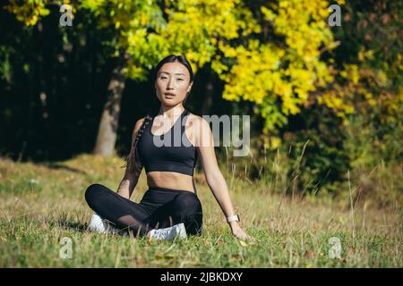 Die schöne asiatische Frau meditiert an einem sonnigen Tag im Wald, die Athletin ruht in der Lotusposition und blickt glücklich und ruhig auf den Kamerataucher Stockfoto