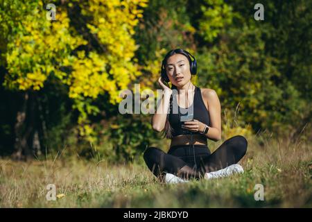 Junge asiatische fit Frau sitzt auf Matte in Lotusposition meditieren entspannend und Musik im Freien hören. Happy Girl weiblich genießt die Natur mit Kopf Stockfoto