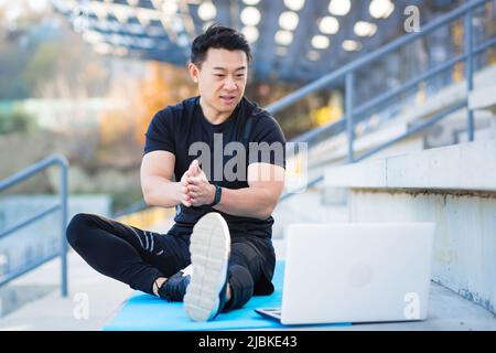 Hübscher asiatischer Fitness-Trainer hält Trainingskurse online mit Videoanruf-Webcam und Laptop. Sitzt draußen in einem modernen Stadion. Pilates Training oder Stockfoto