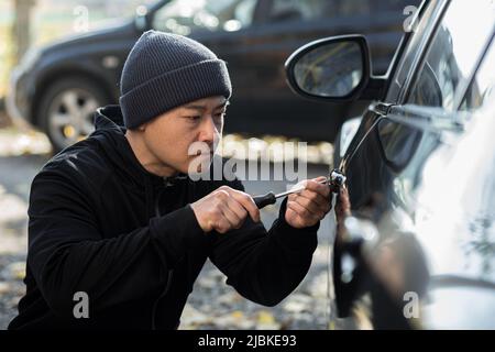 Männlicher asiatischer Dieb stiehlt Auto bricht Türschloss mit mechanischem Schraubendreher-Werkzeug Stockfoto