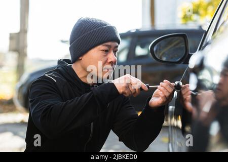 Männlicher asiatischer Dieb stiehlt Auto bricht Türschloss mit mechanischem Schraubendreher-Werkzeug Stockfoto