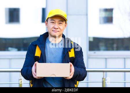 Walking Kurier in schützende medizinische Maske auf Gesicht Lieferung Mann Asian lächelnd auf der Suche nach Lieferadresse am Telefon, hat großen gelben Rucksack für Lebensmittel Stockfoto