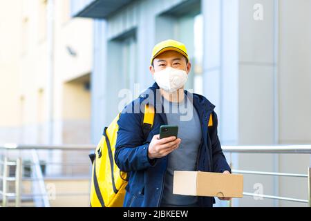 Walking Kurier in schützende medizinische Maske auf Gesicht Lieferung Mann Asian lächelnd auf der Suche nach Lieferadresse am Telefon, hat großen gelben Rucksack für Lebensmittel Stockfoto