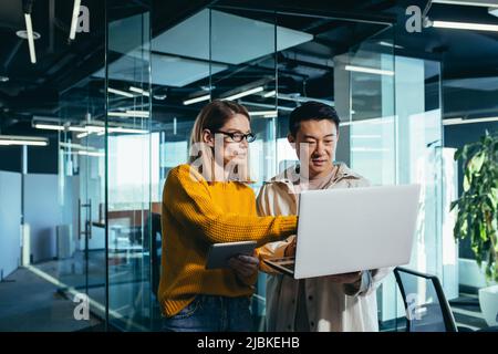 Geschäftstreffen von zwei Mitarbeitern, asiatischen und weiblichen Kollegen, diskutieren das Projekt, arbeiten in einem modernen Büro, Blick auf einen Laptop-Bildschirm, Diskus Stockfoto