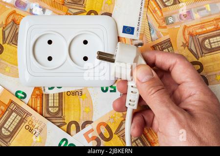 Männliche Hand steckt Stecker in der Steckdose über Geld Hintergrund. Stromkosten und teures Energiekonzept Stockfoto