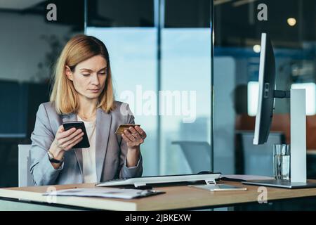 Seriöse Geschäftsfrau Bankkauffrau führt Bankgeschäfte mit einer Kreditkarte und einer Anwendung am Telefon durch, arbeitet in einer modernen Bankfiliale Stockfoto