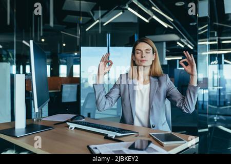Erfolgreiche Geschäftsfrau in Geschäftskleidung, meditierend bei der Arbeit in einem modernen Büro am Computer, versuchend, Ruhe und Balance zu halten Stockfoto