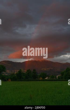 leuchtendes Abendrot mit Regenboden nach dem Sonnenuntergang in Dornbirn, Vorarlberg, Österreich. rote und graublaue Wolken über den grünen Wiesen Stockfoto