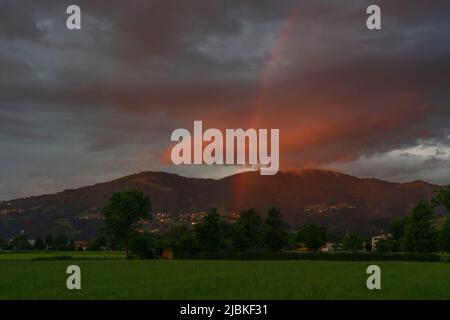 leuchtendes Abendrot mit Regenboden nach dem Sonnenuntergang in Dornbirn, Vorarlberg, Österreich. rote und graublaue Wolken über den grünen Wiesen Stockfoto