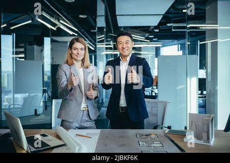 Zwei Architektenteams, die die Kamera betrachten und sich über das abgeschlossene Projekt freuen, halten den Daumen hoch Stockfoto