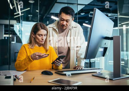 Asiatische Männer und Frauen, Mitarbeiter überweisen per Telefonanwendung und Bankkreditkarte, Kollegen arbeiten in einem modernen Büro zusammen Stockfoto