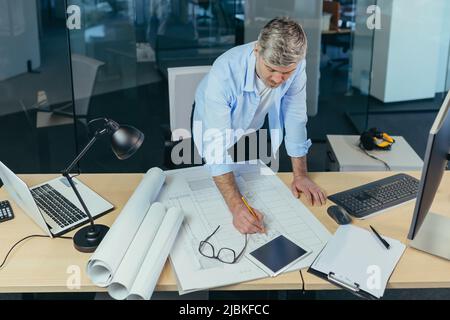 Ein erfahrener Architekt, der am Schreibtisch an der Zeichnung arbeitet, nimmt letzte Änderungen vor der Übergabe des Objekts vor, ein Mann in einem modernen Büro Stockfoto