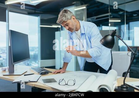 Ein erfahrener Architekt, der am Schreibtisch an der Zeichnung arbeitet, nimmt letzte Änderungen vor der Übergabe des Objekts vor, ein Mann in einem modernen Büro Stockfoto