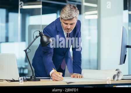 Ein erfahrener Architekt, der am Schreibtisch an der Zeichnung arbeitet, nimmt letzte Änderungen vor der Übergabe des Objekts vor, ein Mann in einem modernen Büro Stockfoto