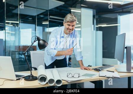 Ein erfahrener Architekt, der am Schreibtisch an der Zeichnung arbeitet, nimmt letzte Änderungen vor der Übergabe des Objekts vor, ein Mann in einem modernen Büro Stockfoto