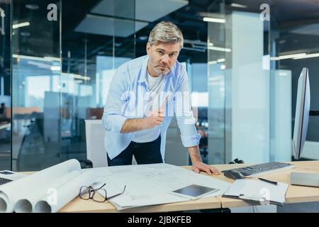 Ein erfahrener Architekt, der am Schreibtisch an der Zeichnung arbeitet, nimmt letzte Änderungen vor der Übergabe des Objekts vor, ein Mann in einem modernen Büro Stockfoto