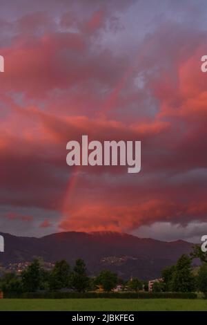 leuchtendes Abendrot mit Regenboden nach dem Sonnenuntergang in Dornbirn, Vorarlberg, Österreich. rote und graublaue Wolken über den grünen Wiesen Stockfoto