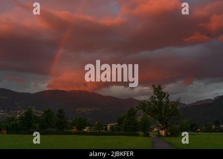 leuchtendes Abendrot mit Regenboden nach dem Sonnenuntergang in Dornbirn, Vorarlberg, Österreich. rote und graublaue Wolken über den grünen Wiesen Stockfoto