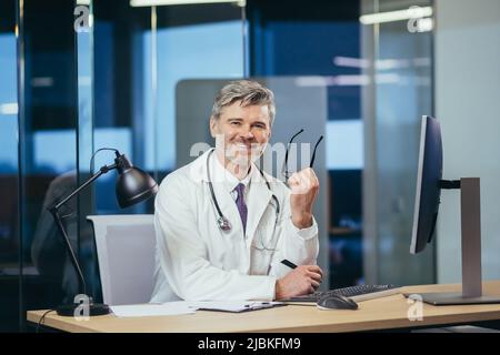 Porträt eines erfahrenen Arztes mit Brille, eines älteren und erfahrenen Mannes, der in die Kamera schaut, eines Arztes, der in einem cli an einem Computer arbeitet Stockfoto