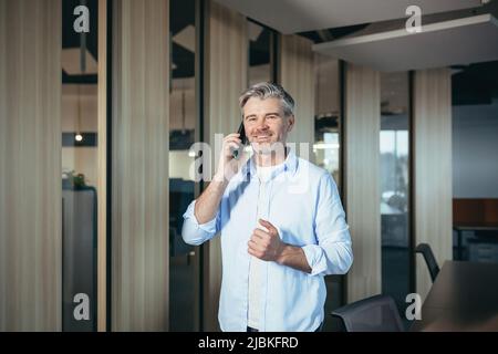 Geschäftsmann, der am Telefon sprach, grauhaariger und erfahrener Mann, der die Kamera ansah und lächelte, Mann bei der Arbeit Stockfoto