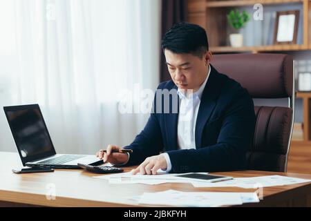 Der asiatische Buchhalter arbeitet im Büro, berechnet den Bericht, der Geldgeber arbeitet auf Papier, verwendet den Laptop und den Rechner Stockfoto