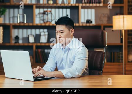 Müde asiatische Geschäftsmann arbeitet spät, in einem klassischen Büro, arbeitet auf einem Laptop, nachdenklich Stockfoto