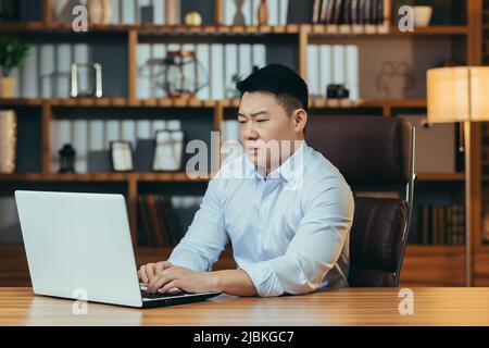 Müde asiatische Geschäftsmann arbeitet spät, in einem klassischen Büro, arbeitet auf einem Laptop, nachdenklich Stockfoto
