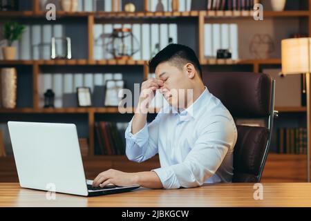 Müde asiatische Geschäftsmann arbeitet spät, in einem klassischen Büro, arbeitet auf einem Laptop, nachdenklich hält seine Hand hinter dem Kopf Stockfoto