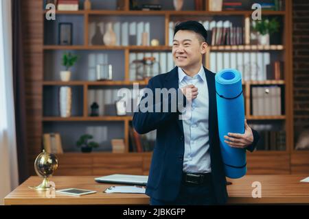 Der Geschäftsmann hat seinen Arbeitstag beendet und bereitet sich auf die Fitness vor, Asian arbeitet in einem klassischen Büro, spät hält der Mann eine Matte für die Fitness Stockfoto