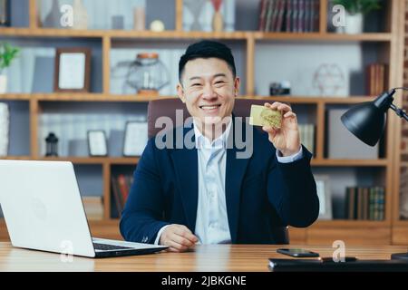 Glücklicher und aufregender asiatischer Bankkaufmann, der die Kamera anschaut und lächelt, im klassischen Büro arbeitet, den Daumen hoch hält und Goldbank-Kredi anbietet Stockfoto